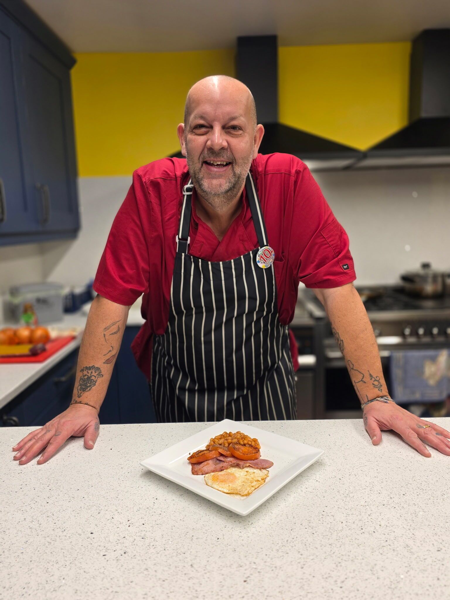 A person in a red shirt and striped apron presenting a plate of food in a kitchen with blue cabinets and a yellow wall.