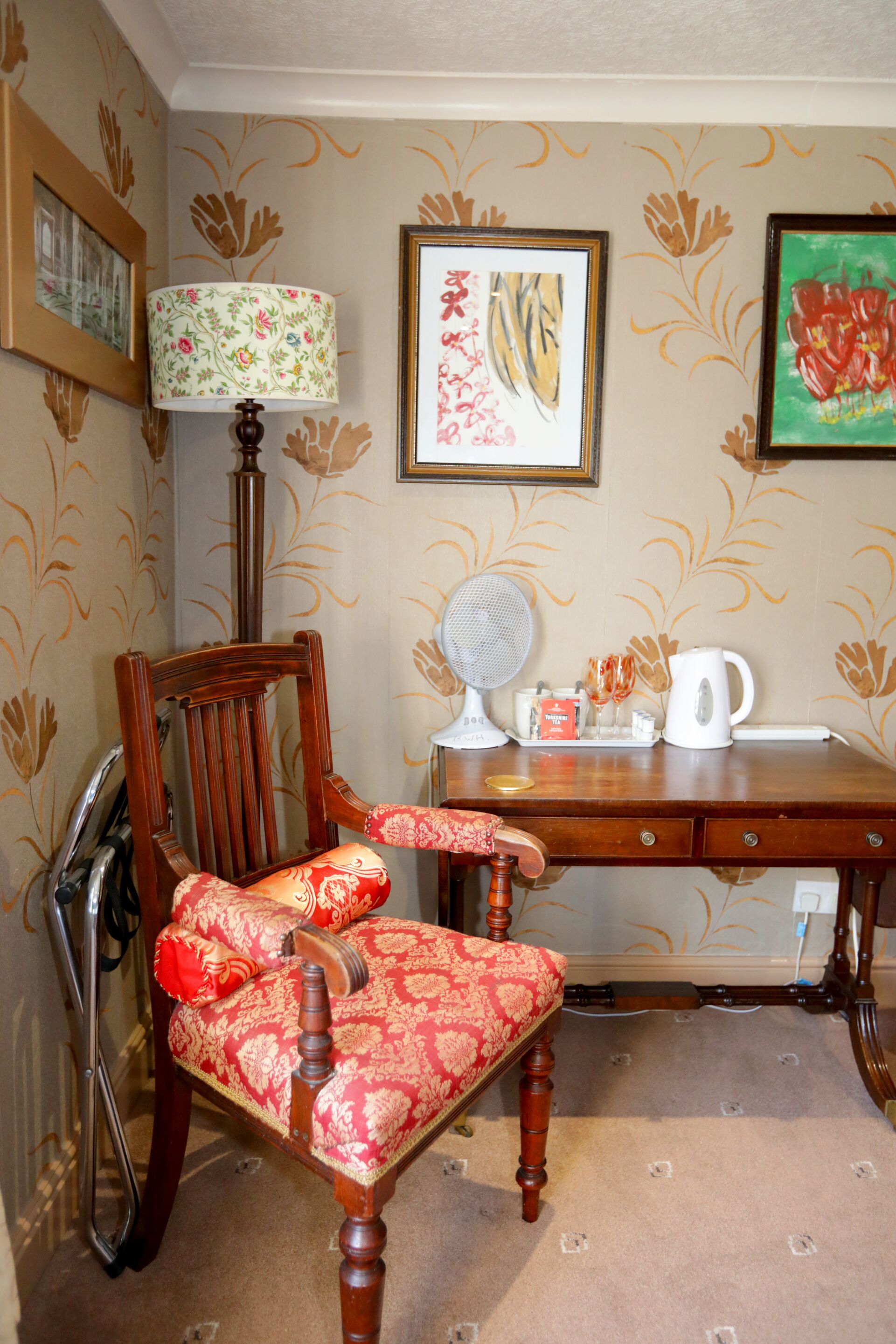 A room with a wooden chair with red upholstery, a wooden desk with a lamp, a framed picture, and a radiator.