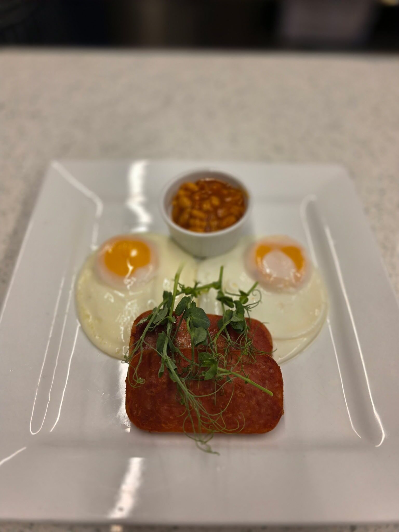 A square plate with two sunny-side-up eggs, a slice of cured meat, and a small bowl of sauce.