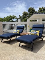Dos sillones de descanso con cojines azules y almohadas con la palabra 'BEACH', sobre una superficie de grava, con una cerca blanca y árboles al fondo.
