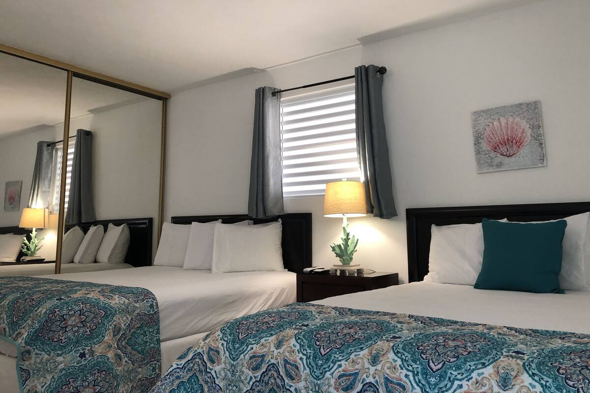 A hotel room with two beds, a large mirror, a window with curtains, a nightstand with a lamp, and a wall art piece.