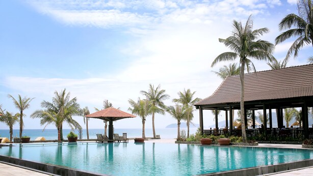 Zona de piscina con palmeras, un espacio de descanso cubierto y vista al mar.