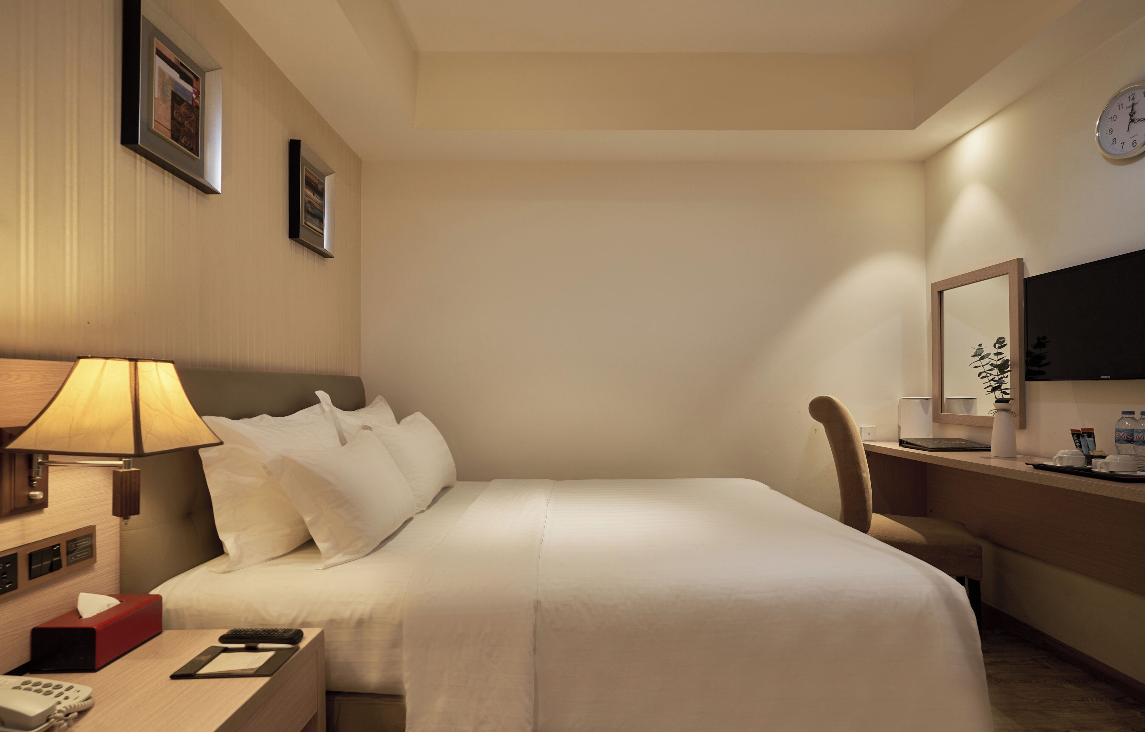 Ein Hotelzimmer mit Bett, Nachttisch, Lampe, Stuhl, Schreibtisch und einer Wanduhr.