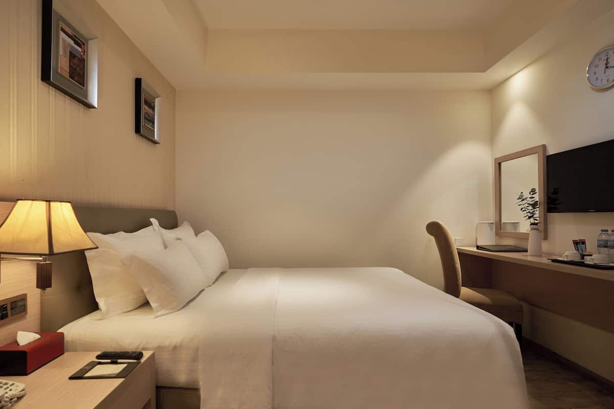 Ein Hotelzimmer mit Bett, Nachttisch, Lampe, Stuhl, Schreibtisch und einer Wanduhr.