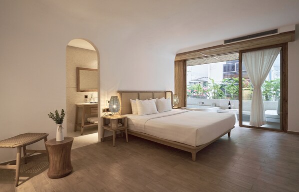 Habitación de hotel con una cama grande, una mesita de noche con lámpara, una pequeña mesa con un jarrón y un ventanal con cortinas.