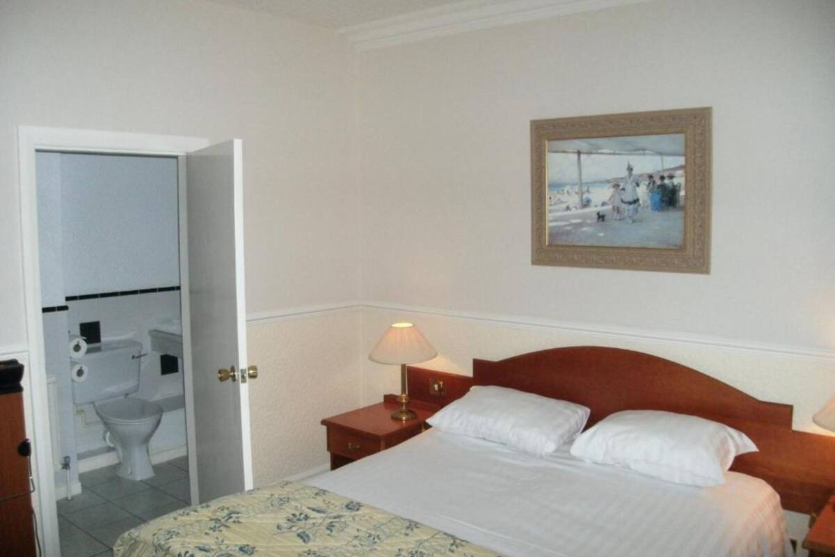 A hotel room with a bed, a bedside table with a lamp, a framed picture on the wall, and an open door leading to a bathroom.