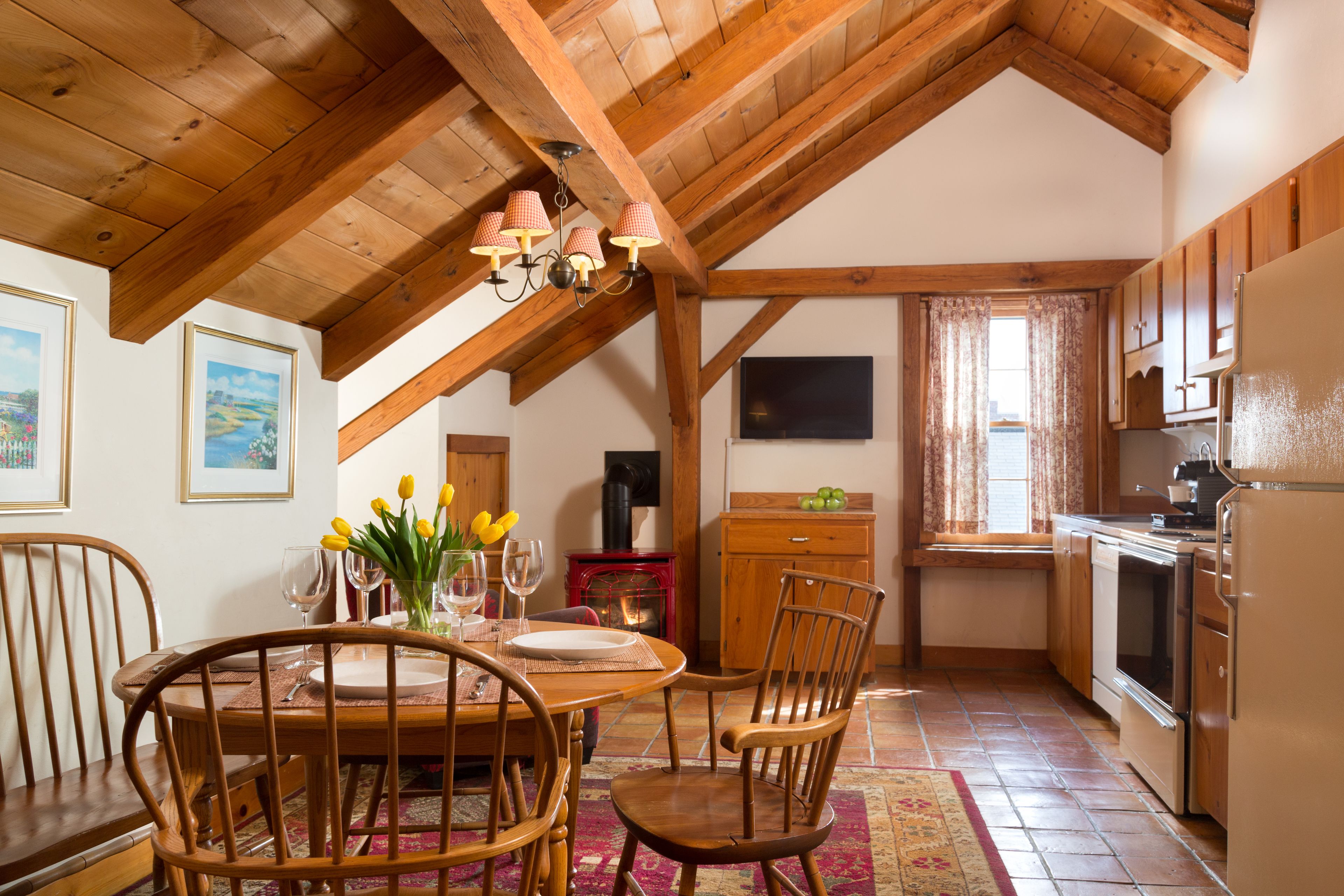 A cozy kitchen with wooden beams, a dining table with chairs, a refrigerator, and a window with curtains.