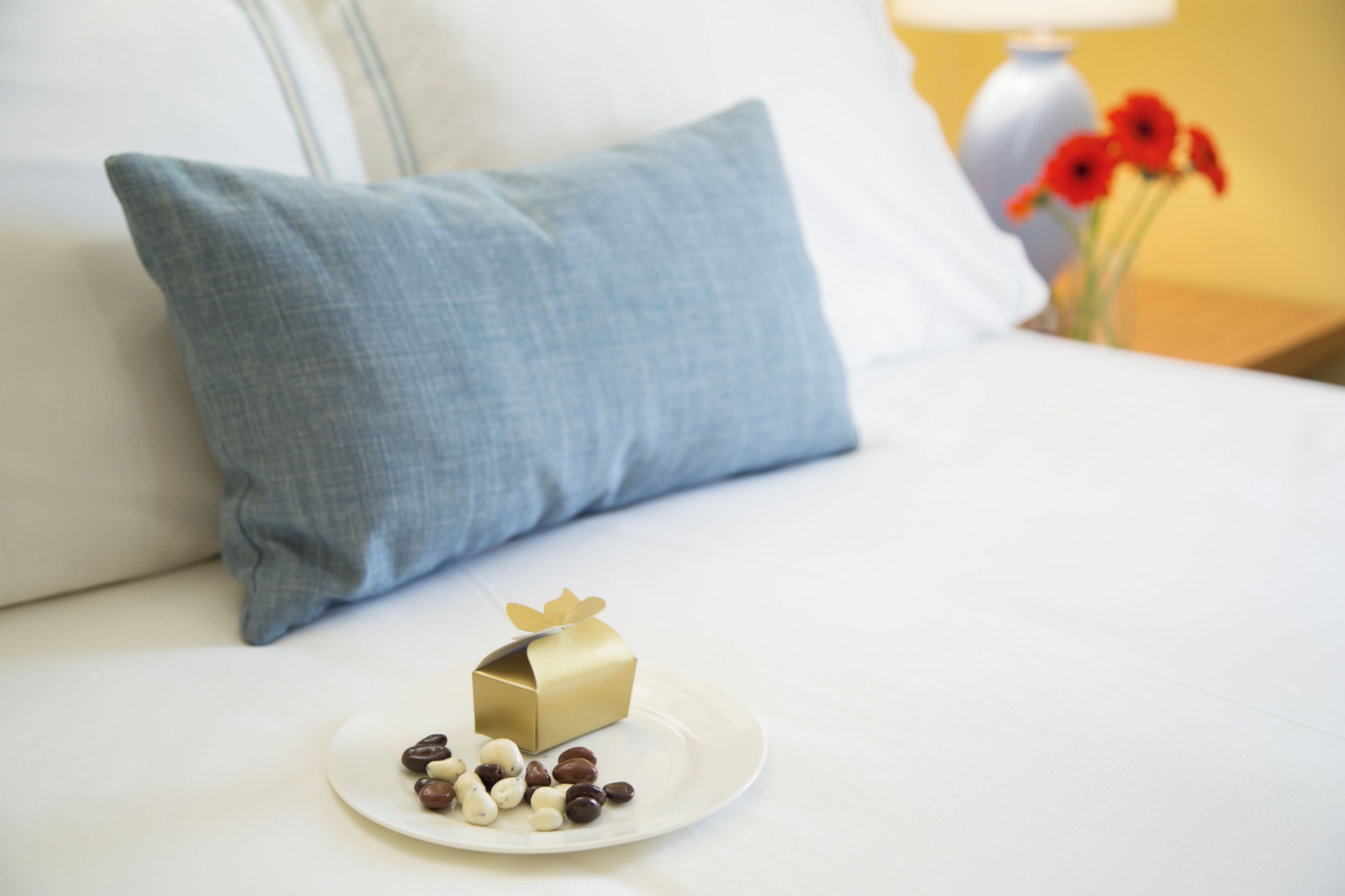 A neatly made bed with a blue pillow, a small gift box, and a plate of chocolates.