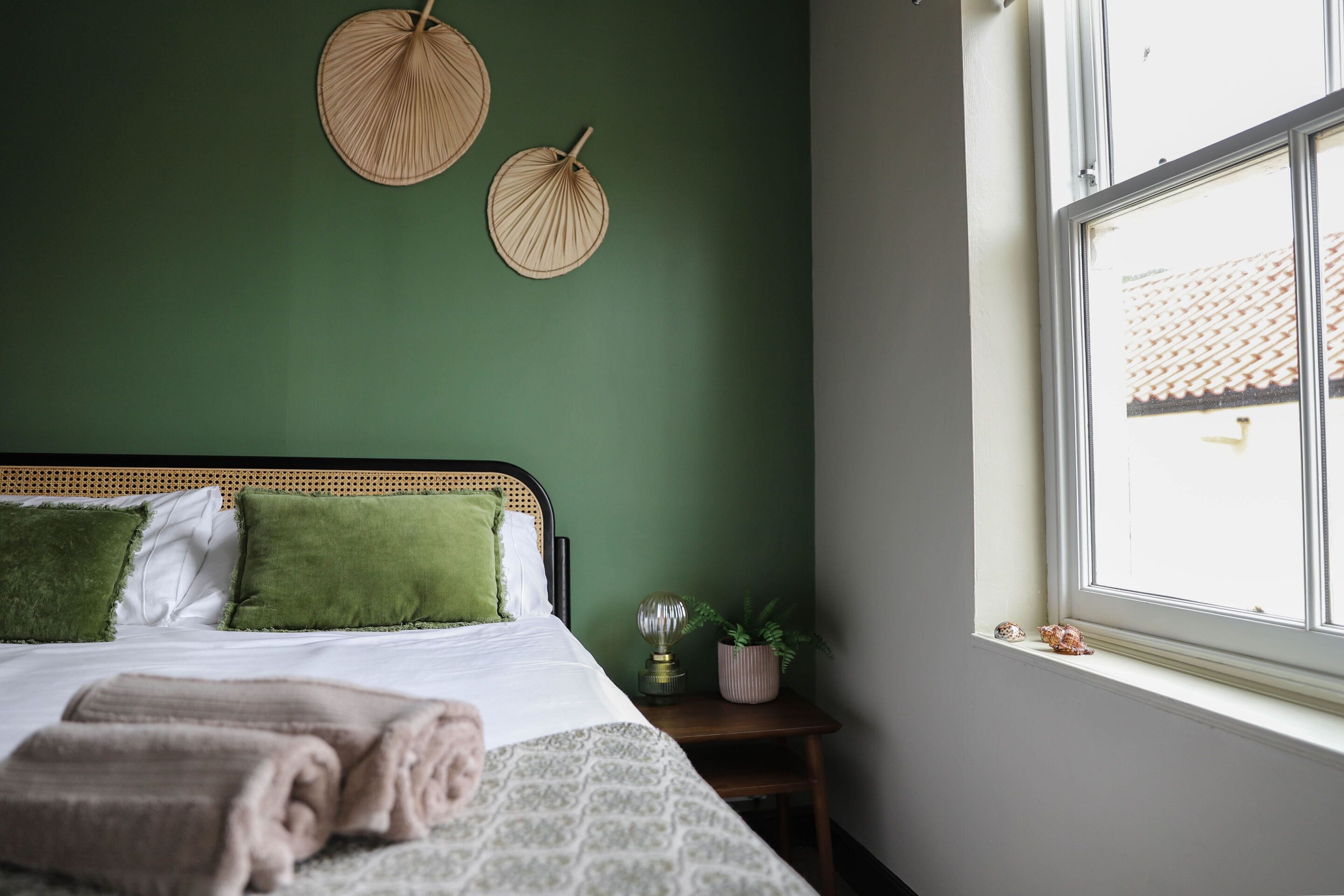 A neatly made bed with green and white bedding, a bedside table with a lamp and a plant, and a window with a view of a building.