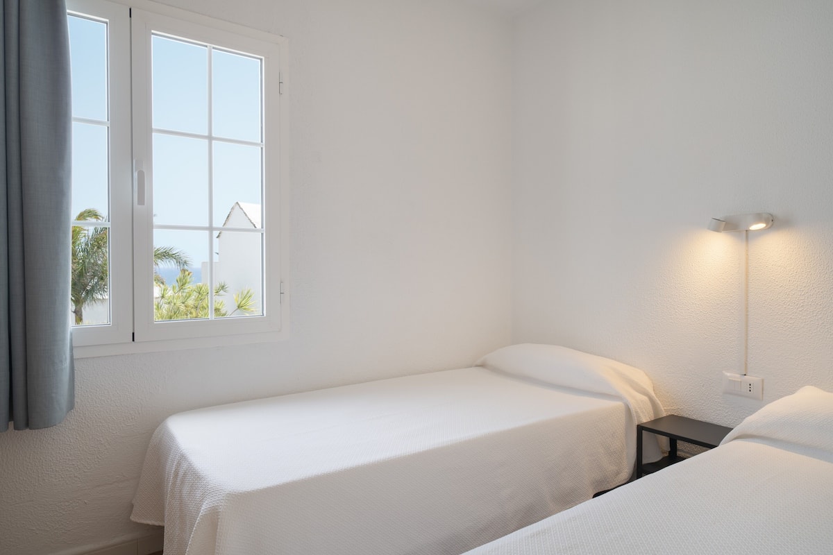 A room with two beds, a window with curtains, and a wall-mounted light fixture.