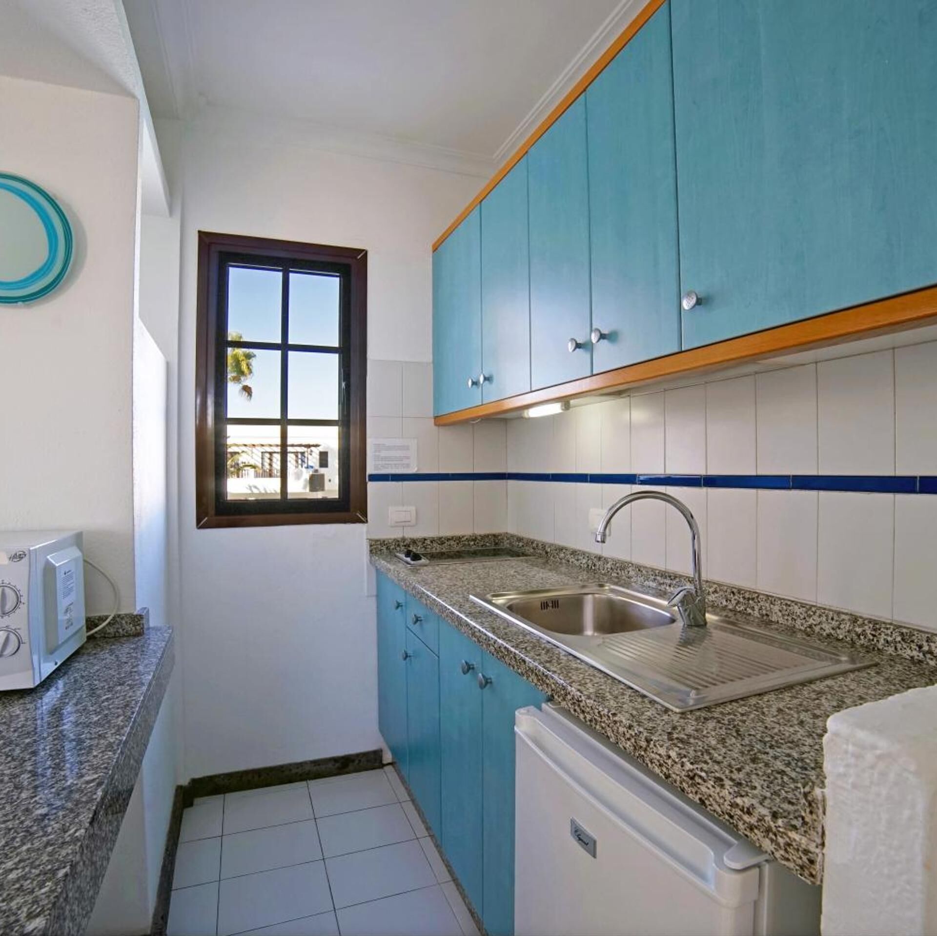 Een kleine keuken met blauwe kastjes, een roestvrijstalen spoelbak en een raam met uitzicht.