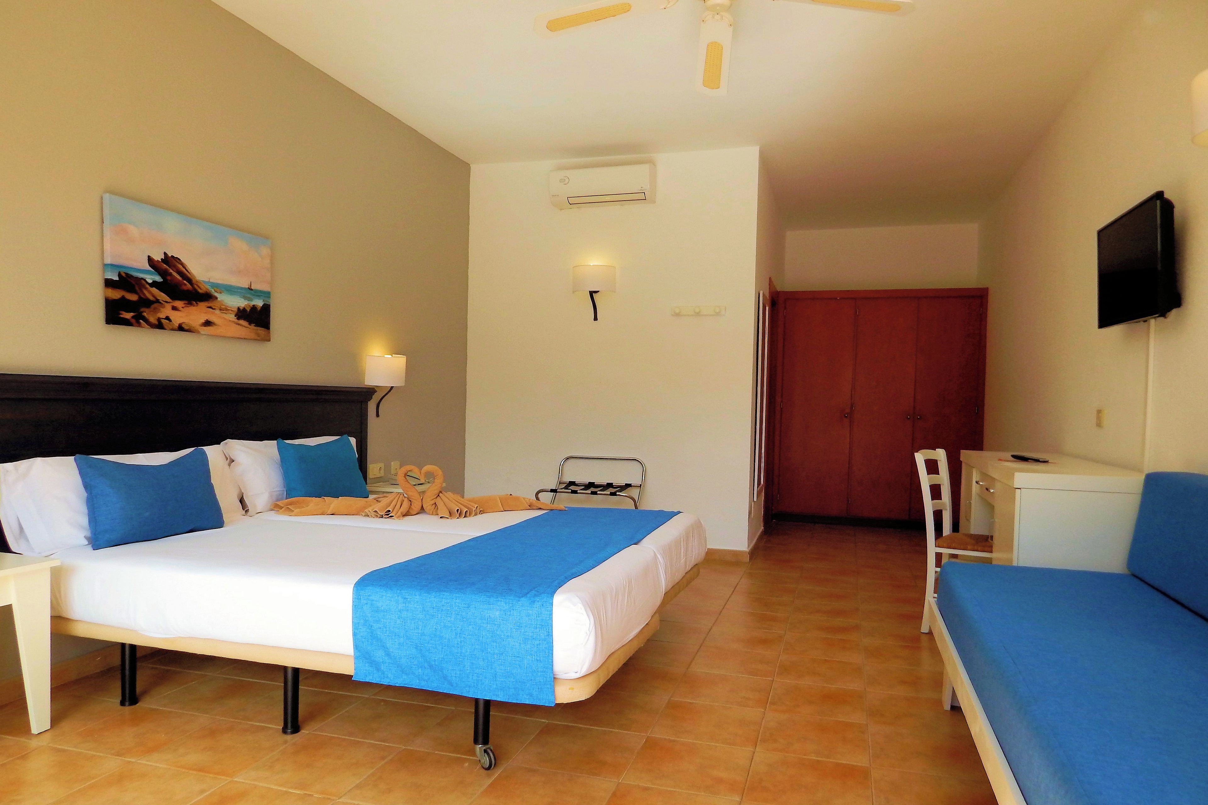 Quarto de hotel com cama, escrivaninha, cadeira, televisão e um quadro na parede.