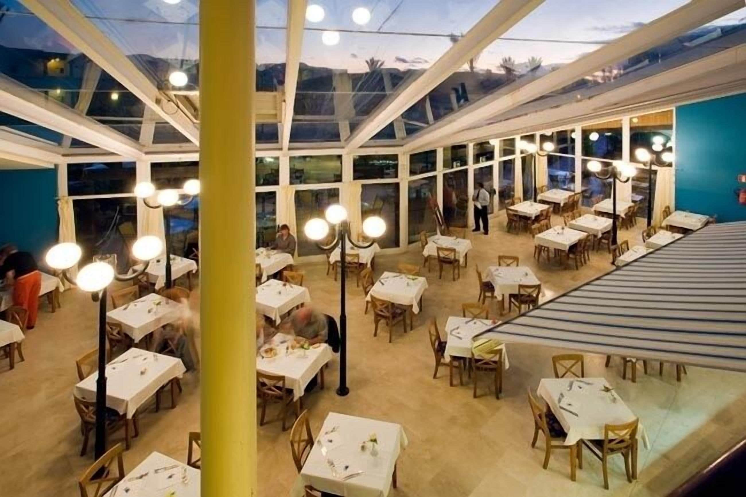 Un restaurante con varias mesas y sillas, ventanas amplias y lámparas colgantes.