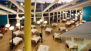 A restaurant with multiple tables and chairs, large windows, and hanging light fixtures. - Monéa Royal Mónica (Yaiza)