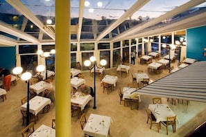 A restaurant with multiple tables and chairs, large windows, and hanging light fixtures. - Monéa Royal Mónica (Yaiza)