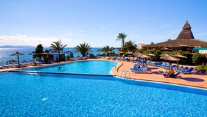 A large outdoor swimming pool with clear blue water, surrounded by lounge chairs and umbrellas, and a view of the sea. - Monéa Royal Mónica (Yaiza)