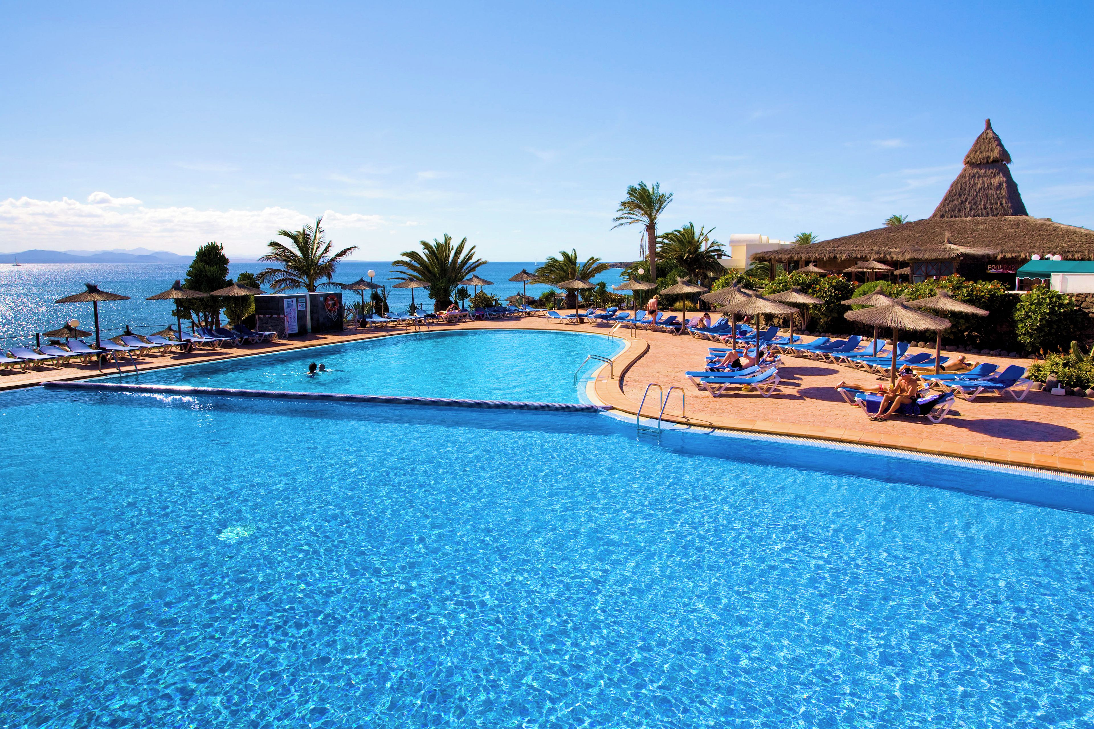 A clear blue pool with lounge chairs and umbrellas, surrounded by palm trees and a view of the sea.