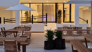 A spacious outdoor patio with wooden tables and chairs, potted plants, and a glass entrance to the building. - Broncemar Beach (Antigua)