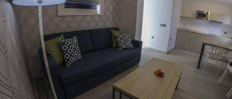 A modern living room with a dark sofa, patterned cushions, a round table, and a floor lamp. A kitchen is visible in the background with a microwave and a dining area.