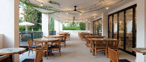 A spacious outdoor dining area with wooden tables and chairs, string lights, and a view of greenery.