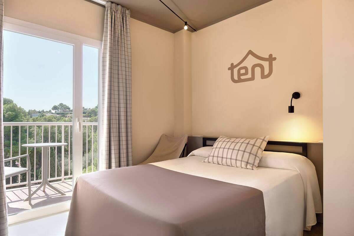 A hotel room with a bed, a nightstand, a wall-mounted light, a balcony with a table and chairs, and a view of greenery outside.