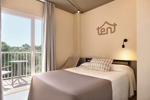 Habitación de hotel con cama, mesita de noche, luz empotrada en la pared, balcón con mesa y sillas, y vista a zonas verdes al exterior.