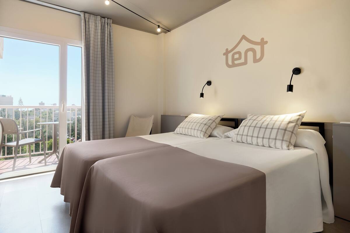 A hotel room with a bed, two bedside tables, a window with curtains, and a balcony with a chair.