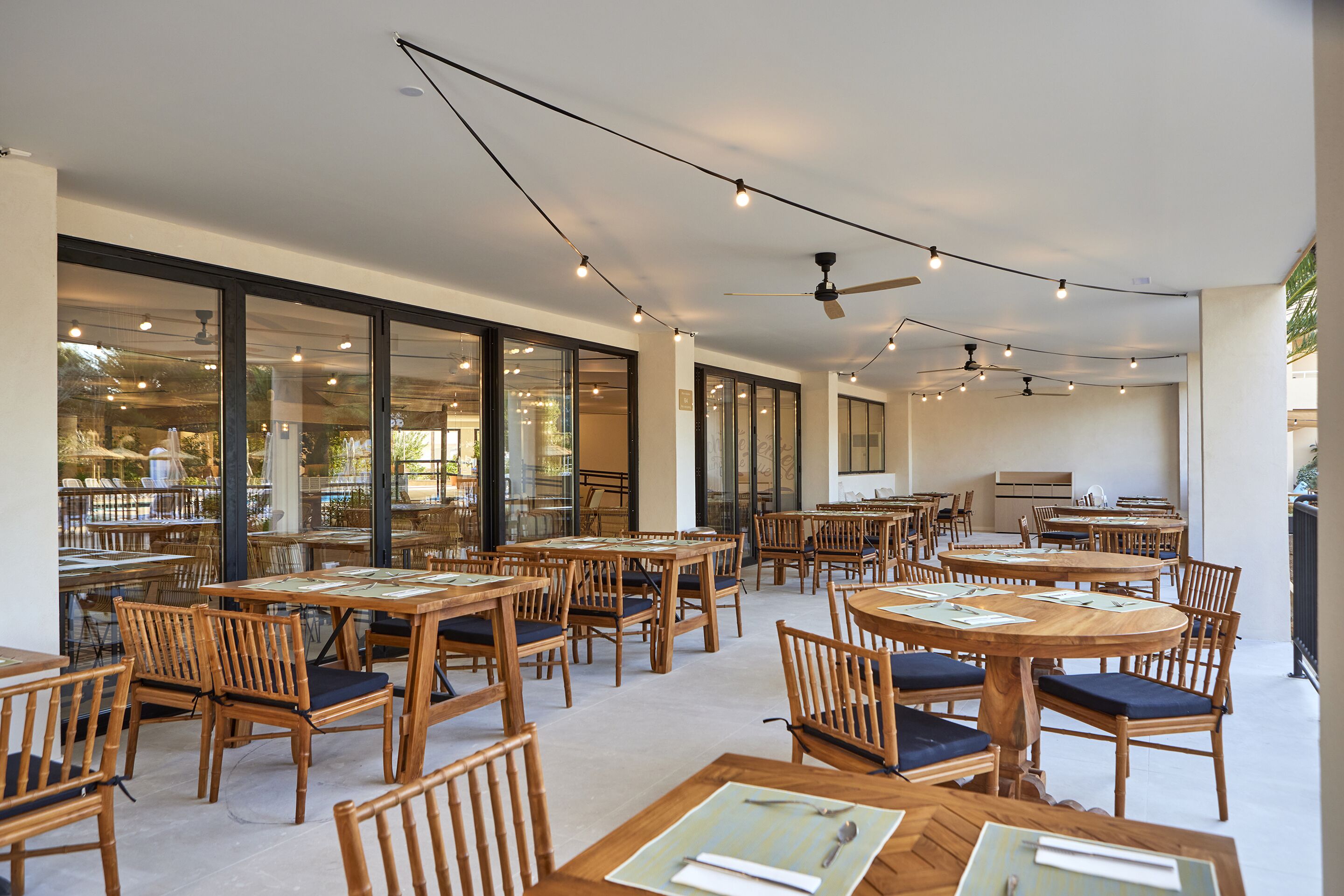 An indoor dining area with wooden tables and chairs, large windows, and ceiling fans.