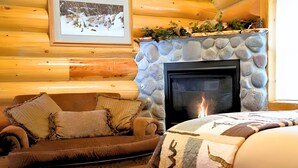 L’intérieur d’un chalet confortable, avec des murs en rondins de bois, une cheminée en pierre, une photographie encadrée accrochée au mur et un canapé en cuir marron agrémenté de coussins décoratifs.