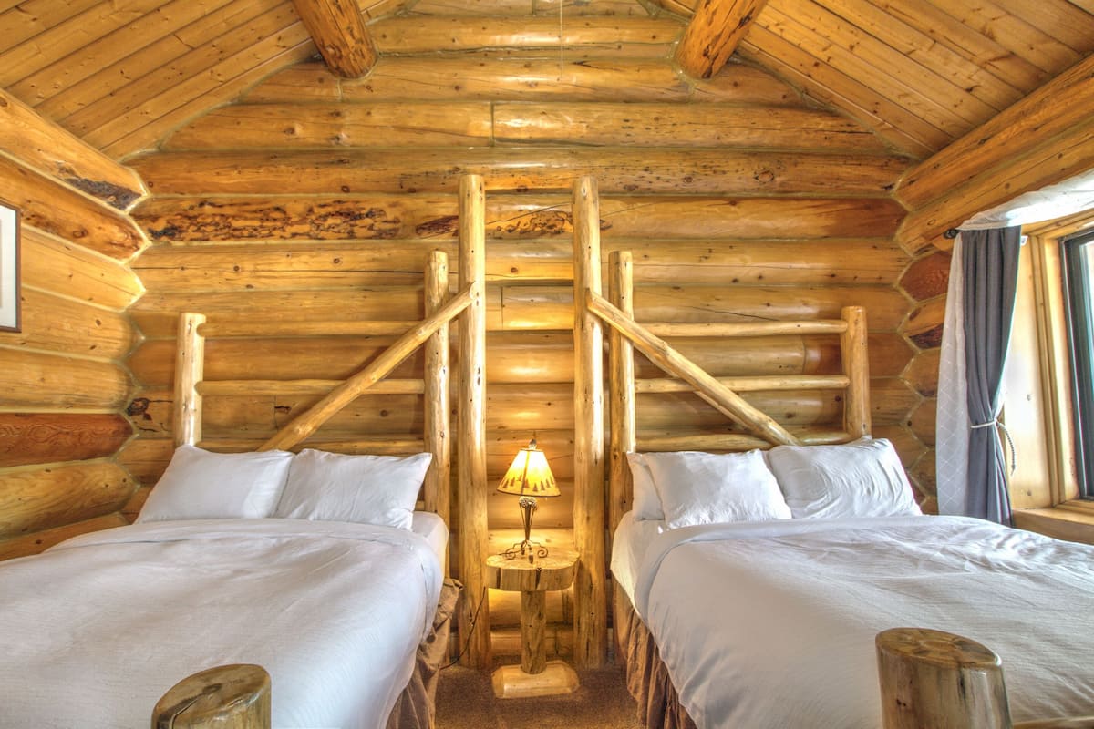 Une chambre aménagée dans un chalet en rondins, avec deux lits, une échelle en bois au centre et une petite table avec une lampe.