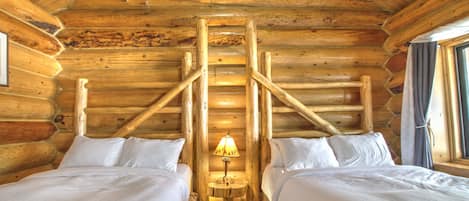 Habitación tipo cabaña de madera con dos camas, una escalera de madera en el centro y una mesita con lámpara.