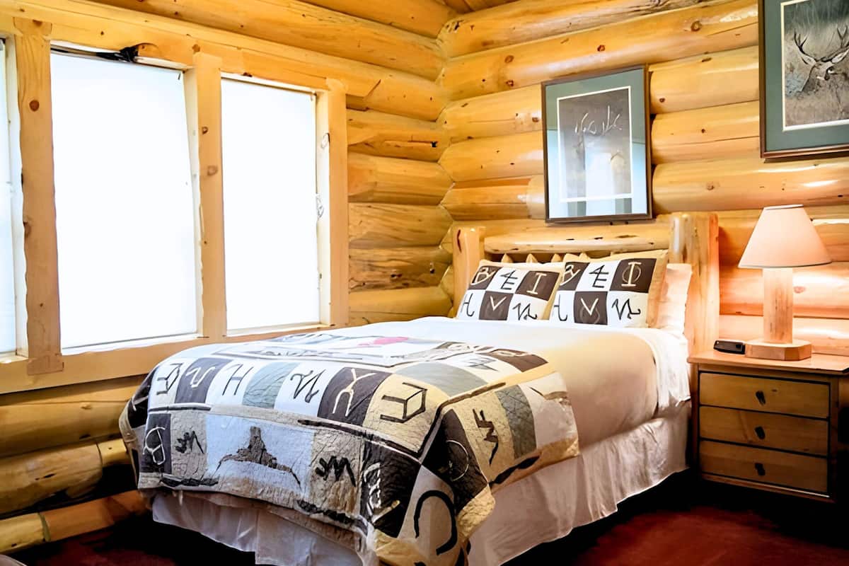 Une chambre située dans une cabane en rondins, avec un lit, une table de chevet, une lampe et deux fenêtres.