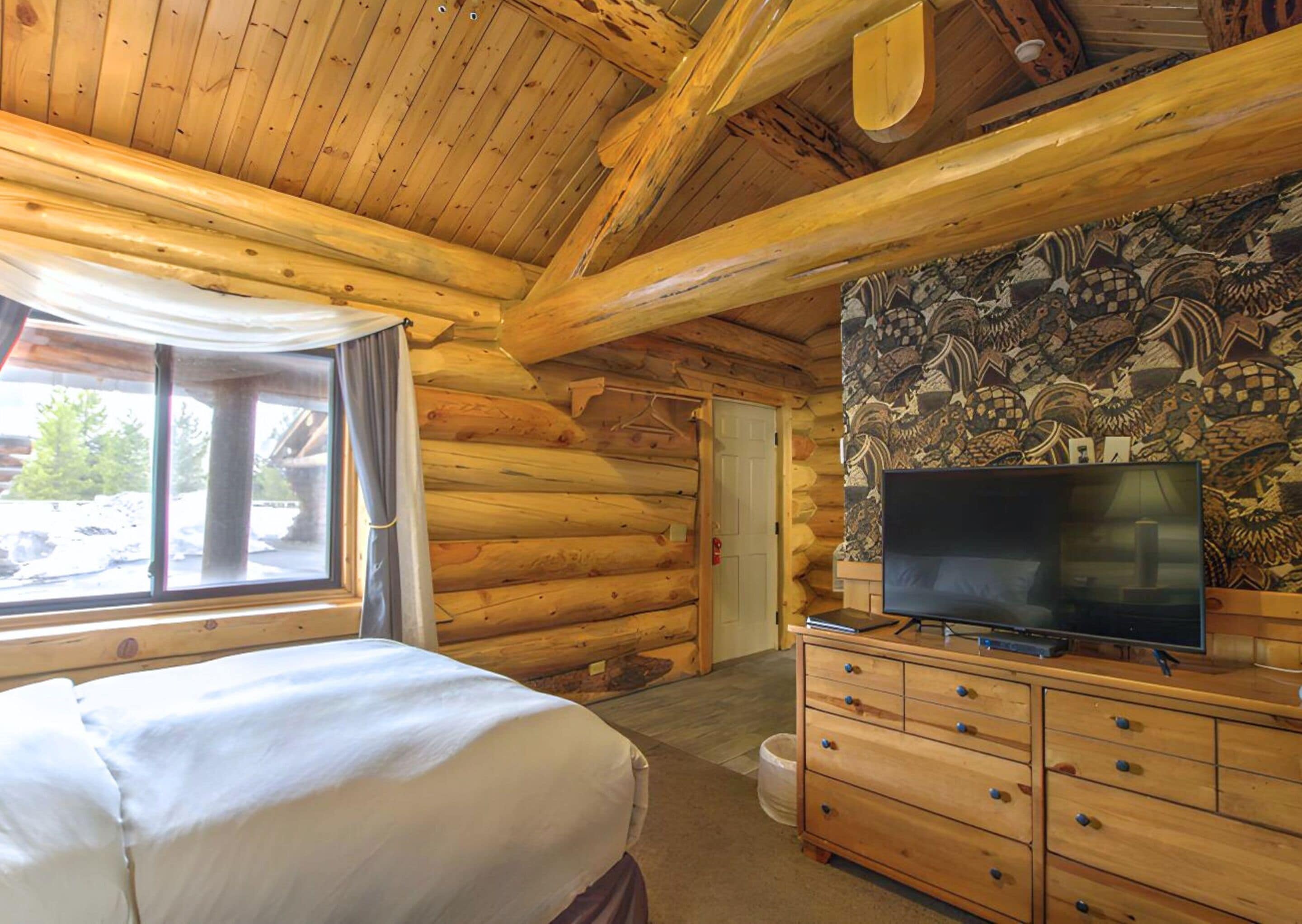 A log cabin room with a bed, a TV on a wooden dresser, and a mural on the wall.