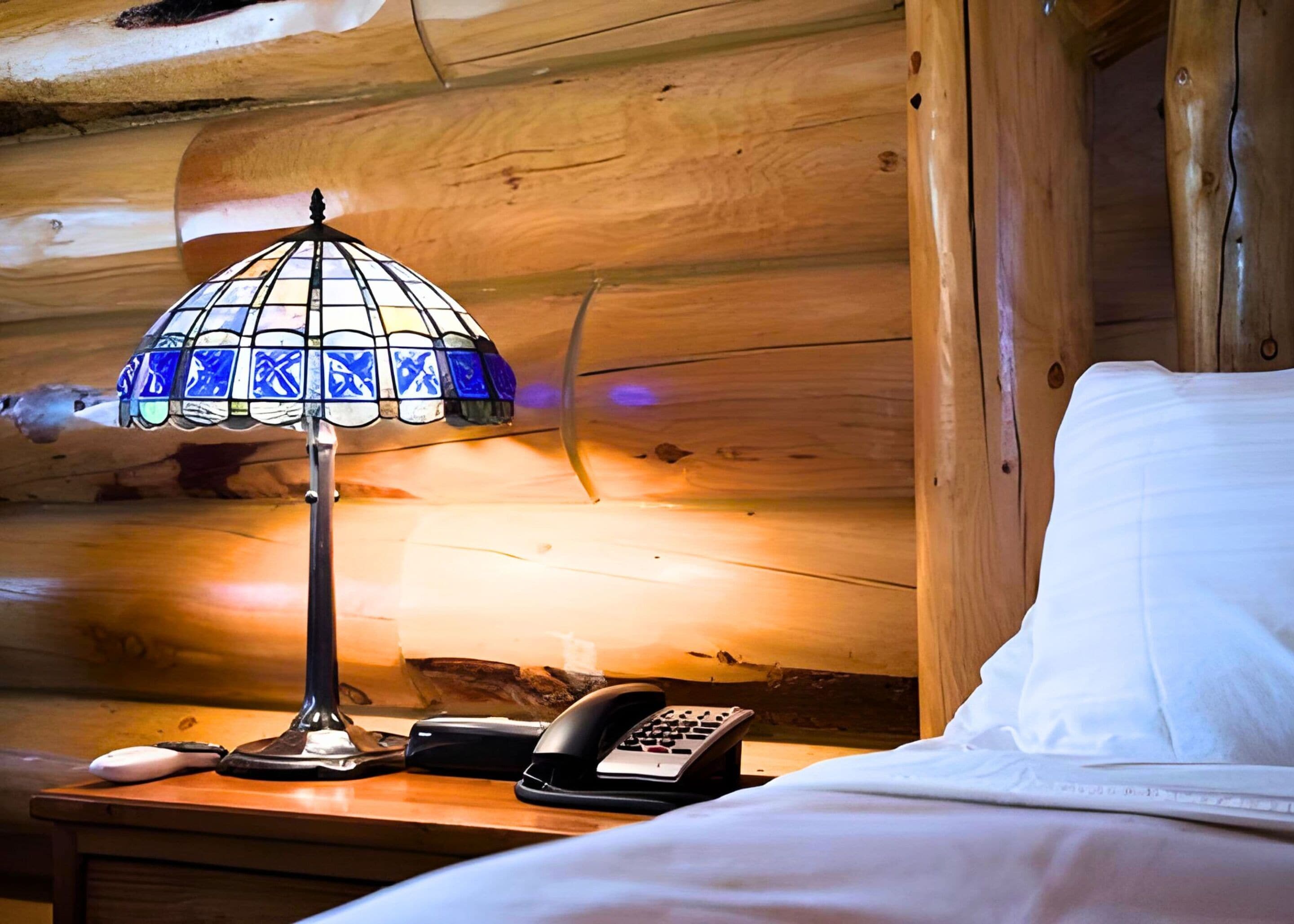 A wooden cabin interior with a bed, a nightstand, a stained glass lamp, and a telephone.