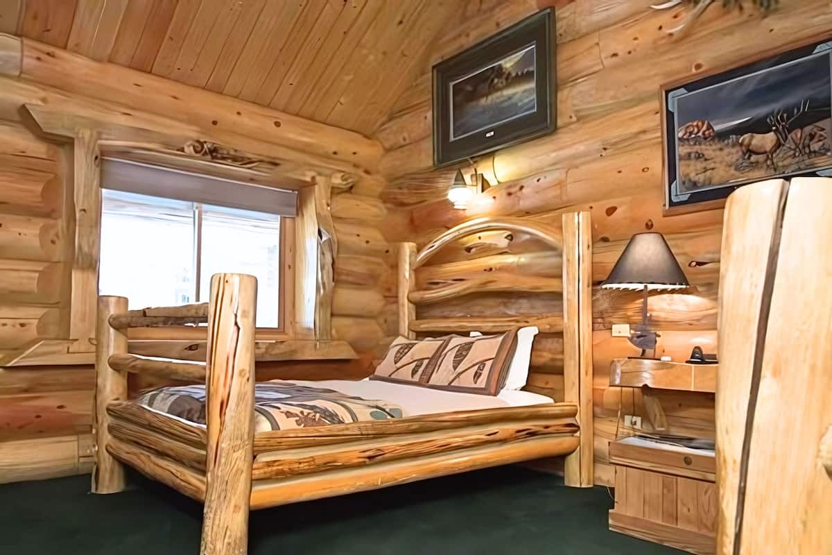 Une chambre dans un chalet en rondins, avec un lit en bois, une fenêtre, une lampe et un tableau encadré au mur.