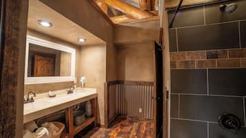 A bathroom with a wooden floor, a sink with a mirror, and a shower area with dark tiles.