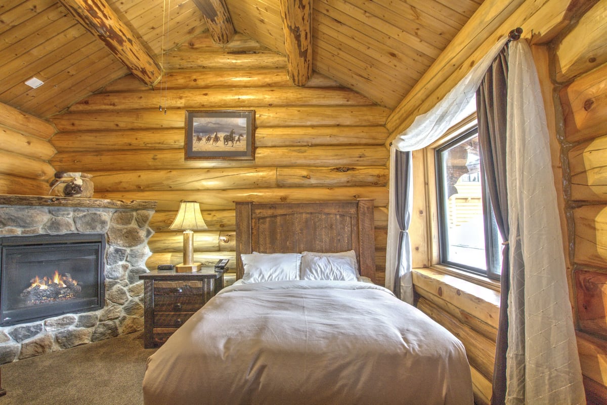 Une chambre située dans une cabane en rondins, avec une cheminée en pierre, un lit recouvert de linge de maison blanc, une table de chevet avec une lampe et une fenêtre avec des rideaux.