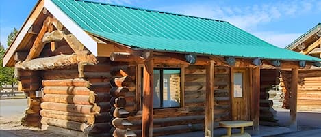 A rustic log cabin with a green metal roof, wooden porch, and a bench.