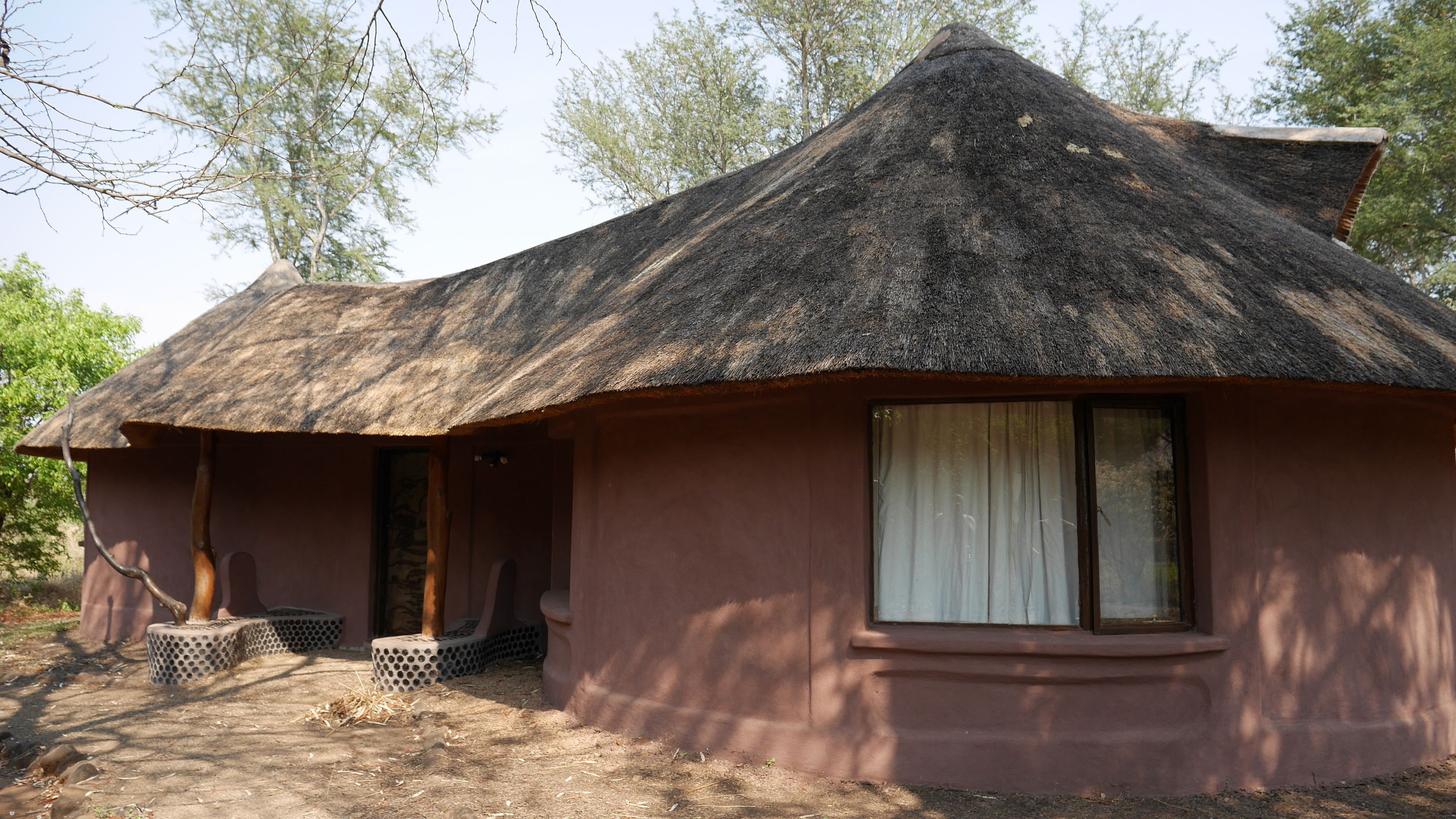 Uma casa com telhado de palha, uma janela e uma porta, cercada por árvores.