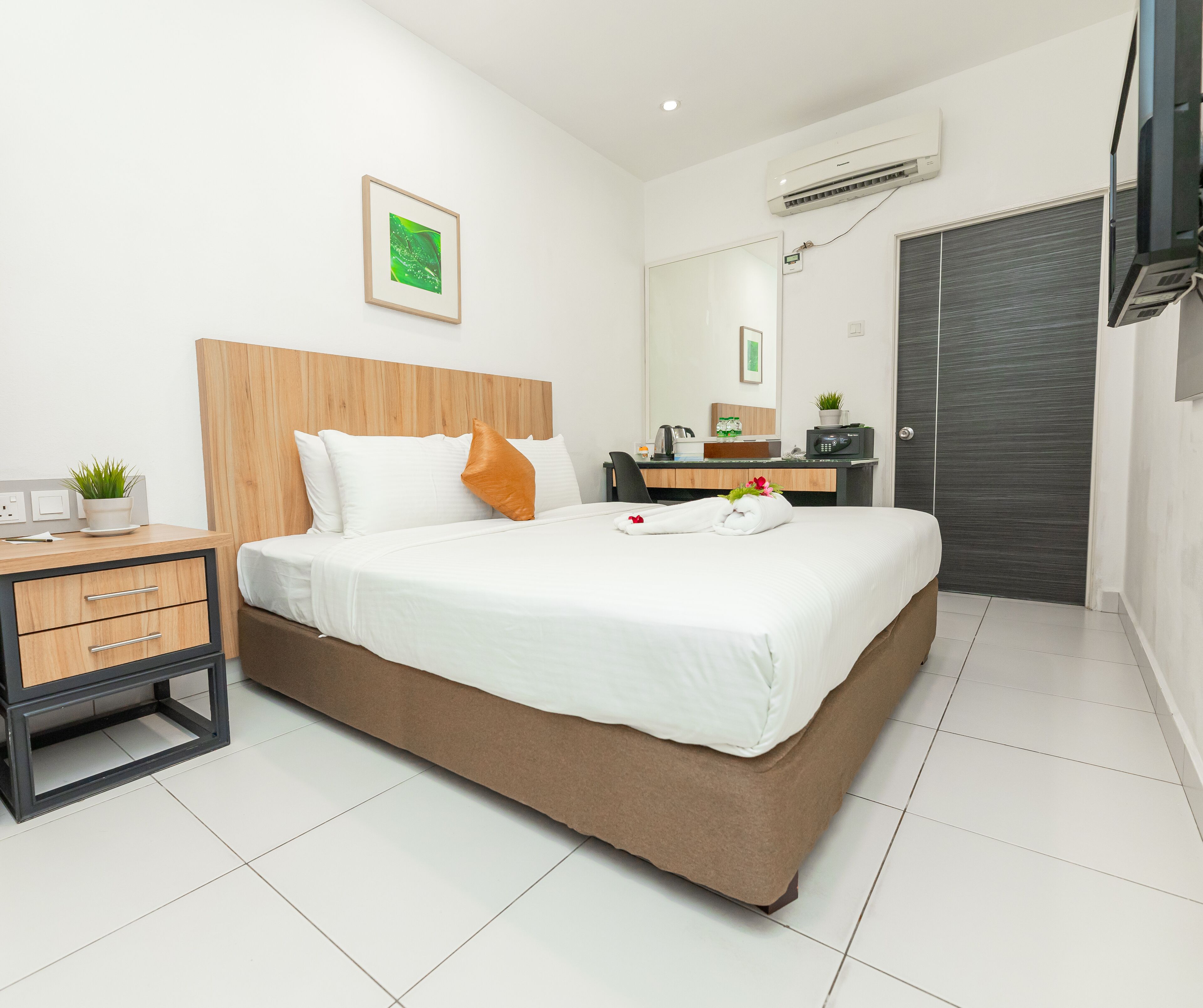 Una habitación de hotel moderna con una cama grande, un cabecero de madera, una mesita de noche con una planta, un escritorio con una silla, una puerta oscura y un equipo de aire acondicionado.