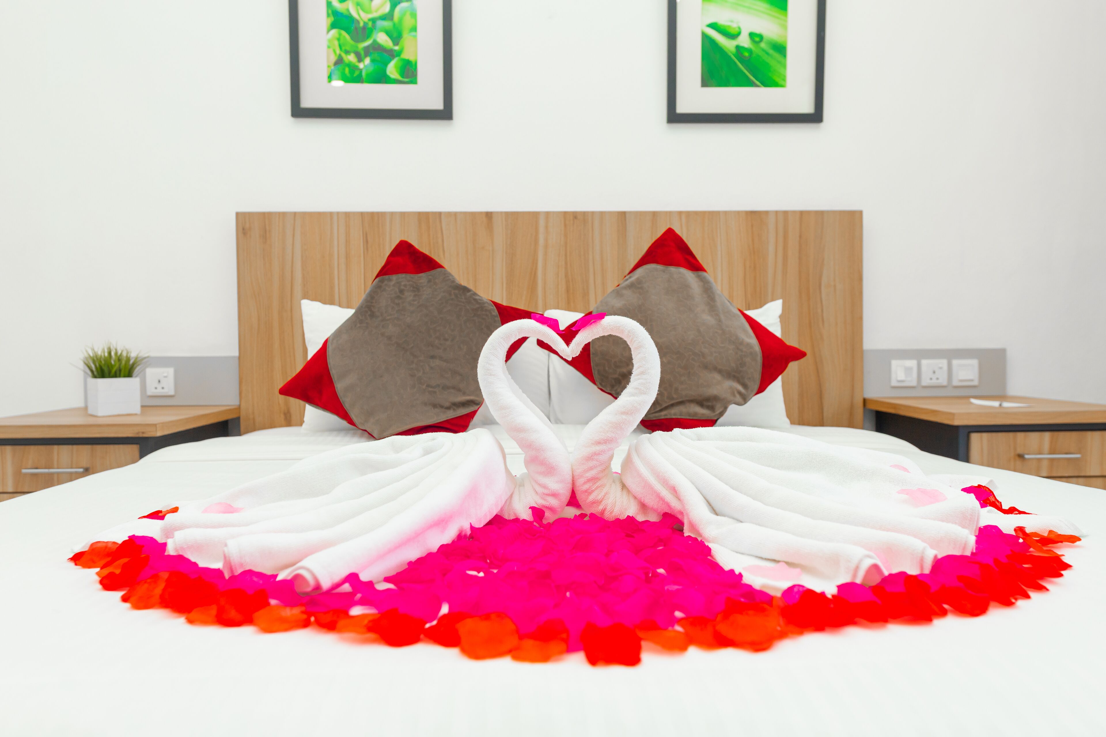 A neatly made bed with a heart-shaped towel arrangement and rose petals, wooden headboard, and two framed pictures on the wall.