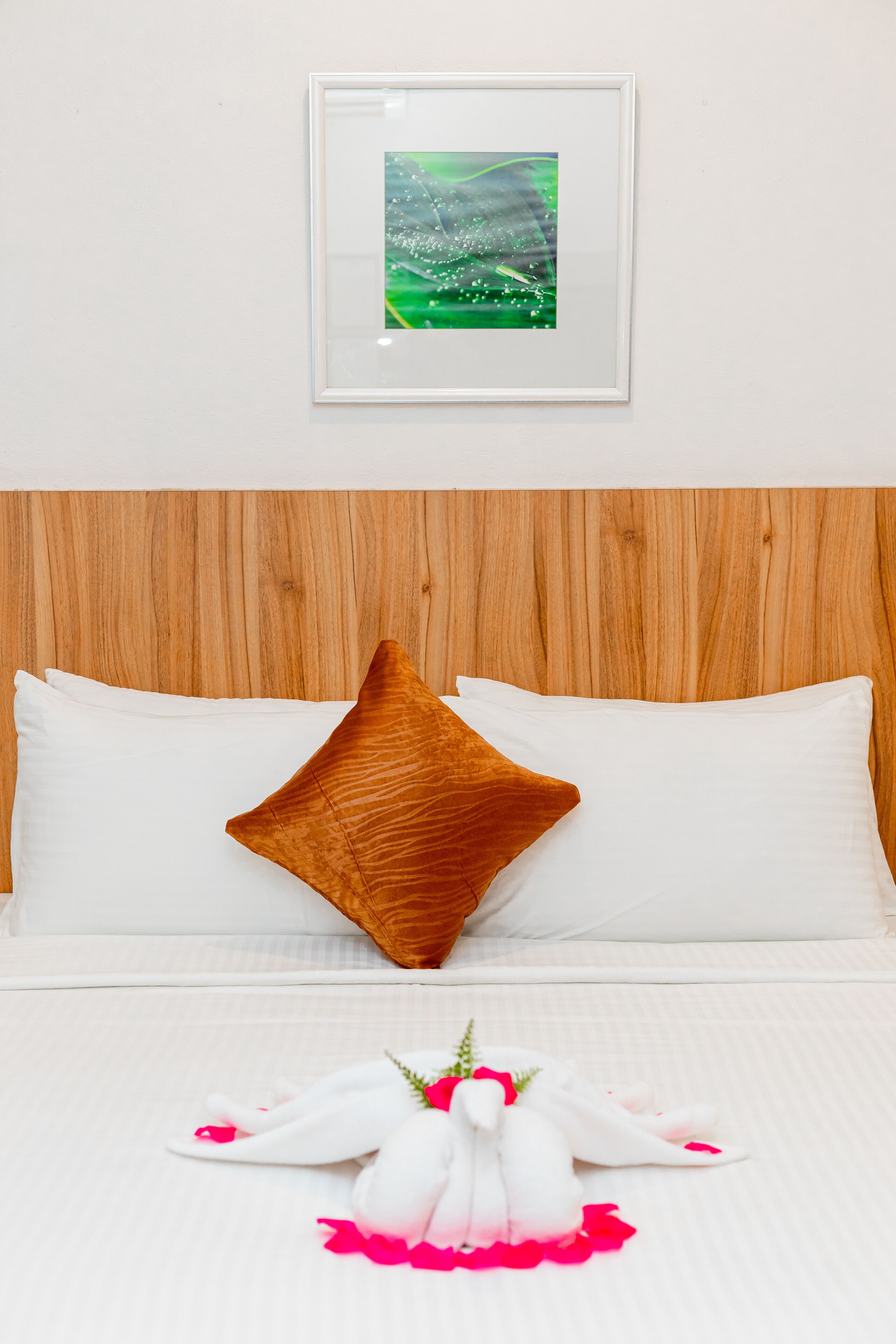 A neatly made bed with white linens and a single brown pillow, a framed picture on the wall, and a decorative towel arrangement on the bed.