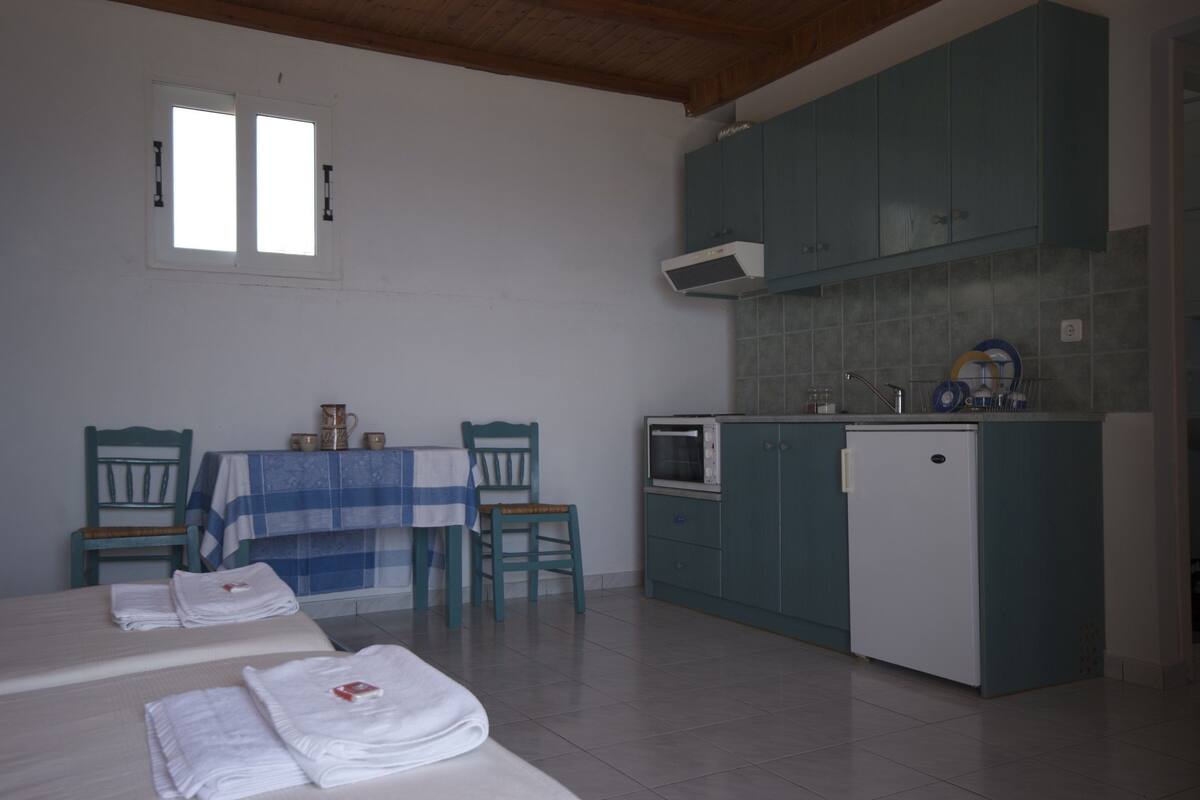 A room with two beds, a small table with a blue and white checkered tablecloth, two chairs, a kitchenette with teal cabinets, a white refrigerator, and a window with blinds.