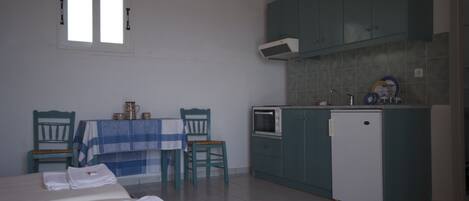 A room with two beds, a small table with a blue and white checkered tablecloth, two chairs, a kitchenette with teal cabinets, a white refrigerator, and a window with blinds.