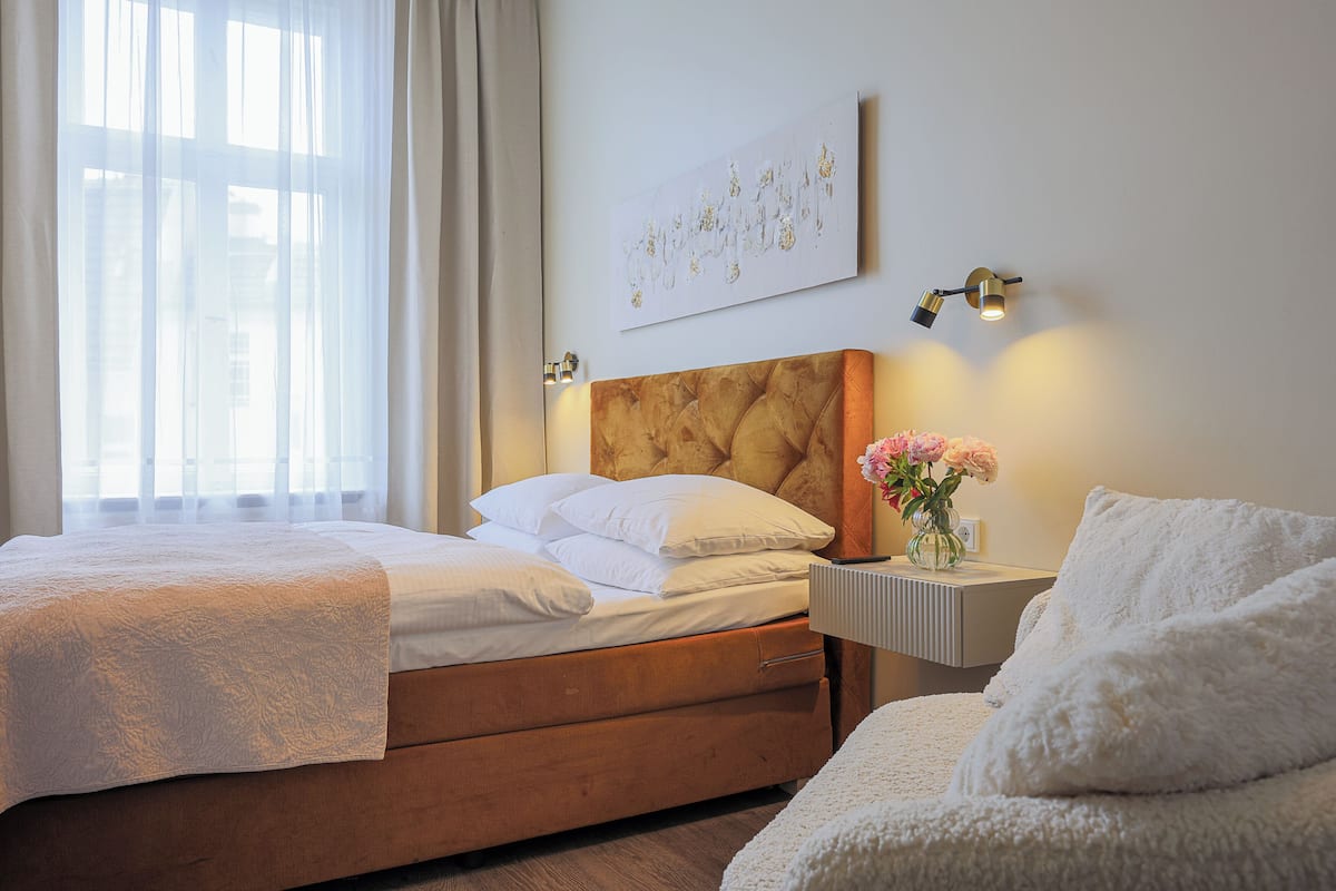 A hotel room with a bed, a bedside table with a lamp, a vase with flowers, and a painting on the wall.