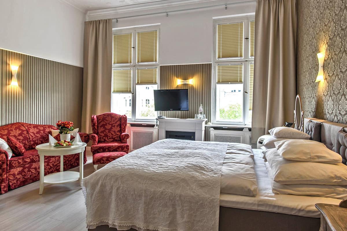 A hotel room with a bed, a TV, a seating area with a chair, and a window with blinds.