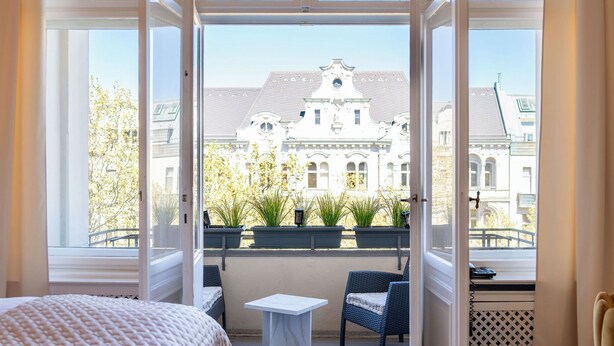Une pièce comprenant un lit, une petite table et des chaises, de grandes fenêtres avec des rideaux, et une vue sur un bâtiment à l’extérieur.