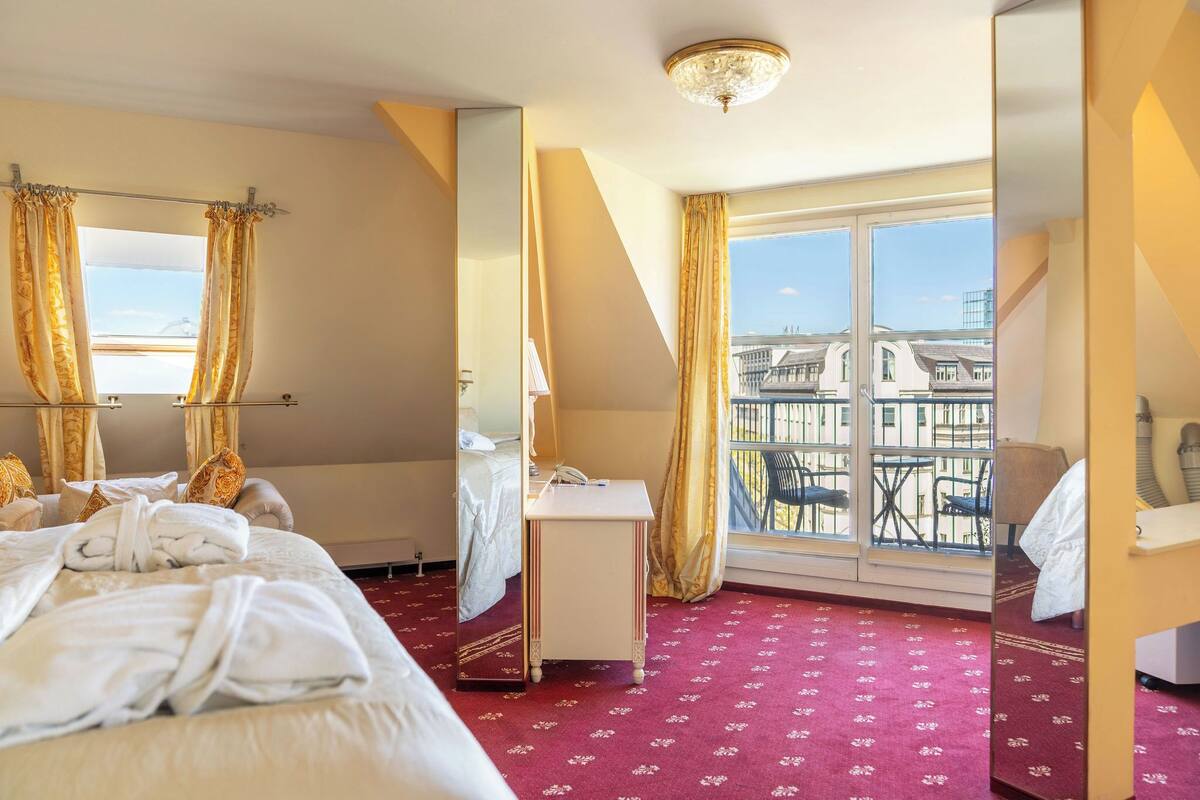 A hotel room with a bed, a desk, a chair, a window with a view of buildings, and a chandelier.