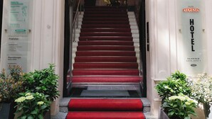 L’entrée d’un hôtel, avec une grande arche, un escalier recouvert d’un tapis rouge et une enseigne indiquant « HÔTEL AUGUSTA ».
