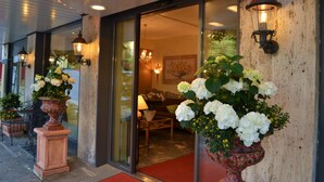 Entrada de hotel con alfombra roja, grandes flores blancas en macetas y faroles en la pared.